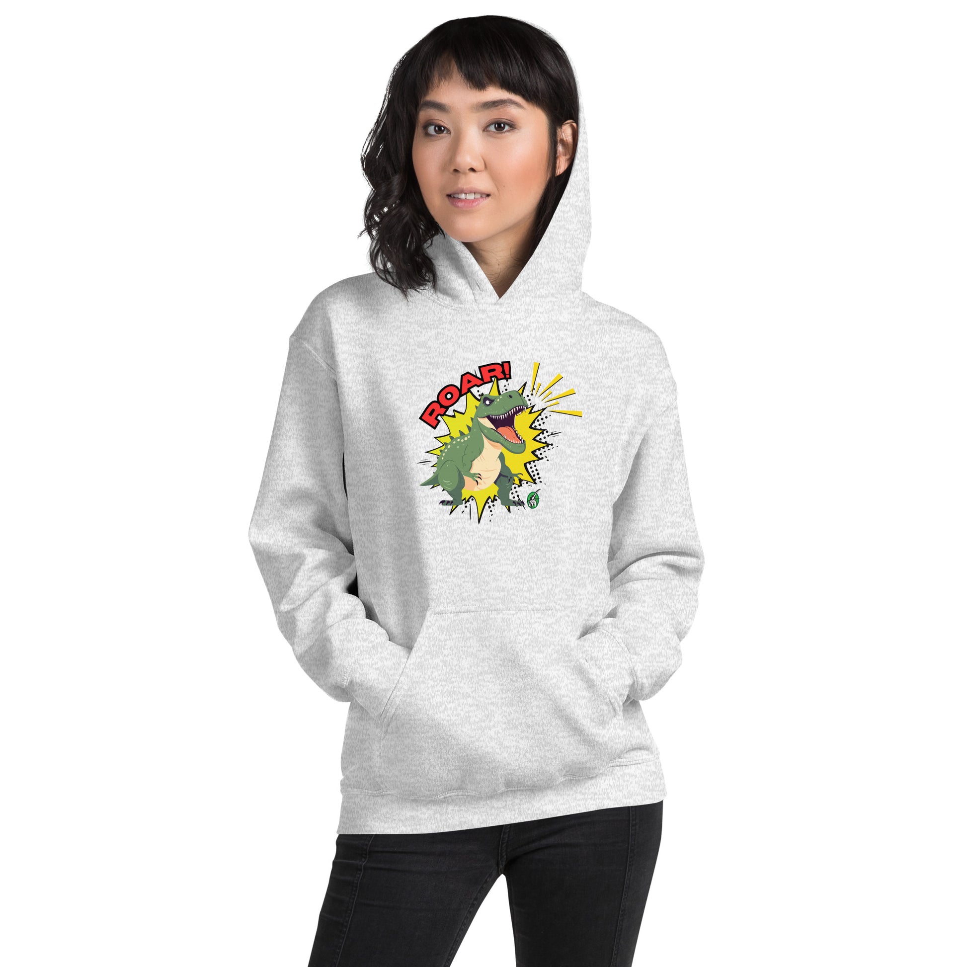 A woman standing wearing an ash hoodie with a printed graphic of a green roaring cartoon dinosaur and the word Roar! Designed by Wizard Prints