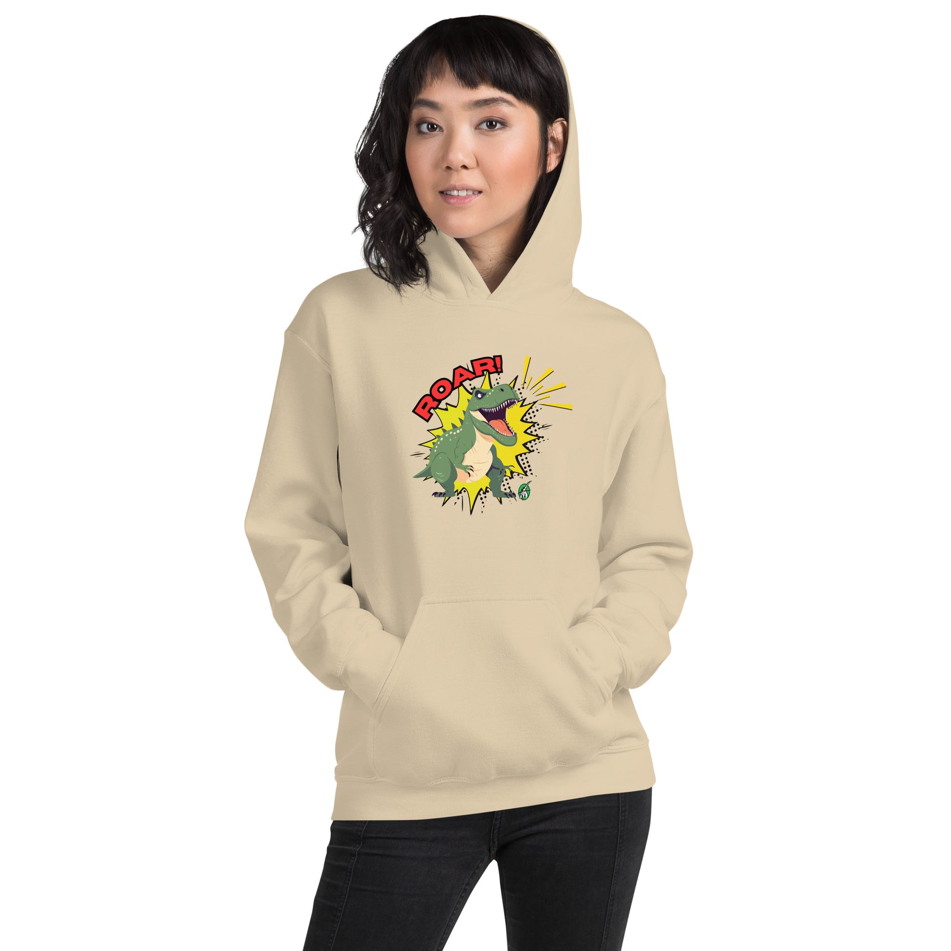 A woman standing wearing a beige hoodie with a printed graphic of a green roaring cartoon dinosaur and the word Roar! Designed by Wizard Prints