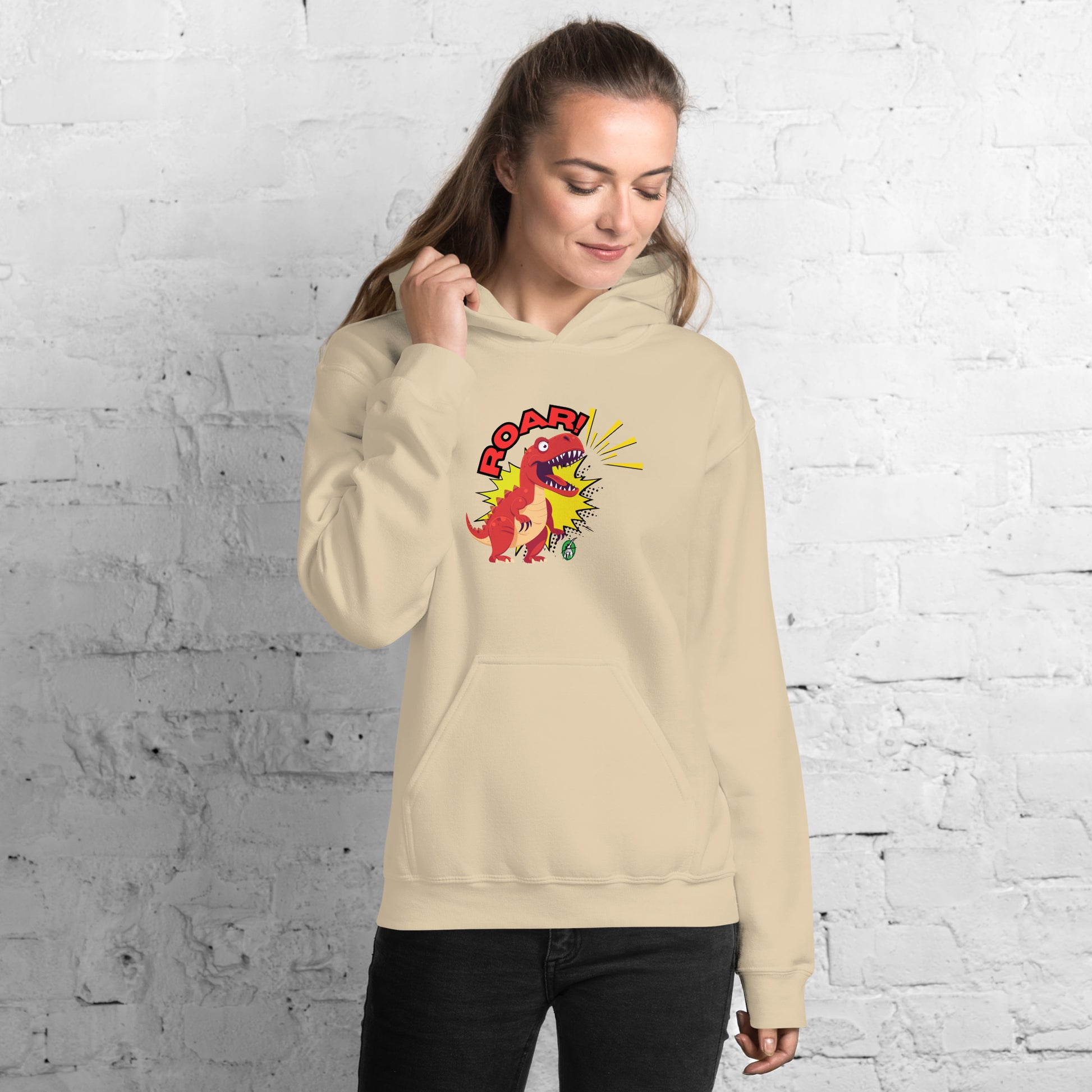 A woman standing wearing a beige hoodie with a printed graphic of a red roaring cartoon dinosaur and the word Roar! Designed by Wizard Prints
