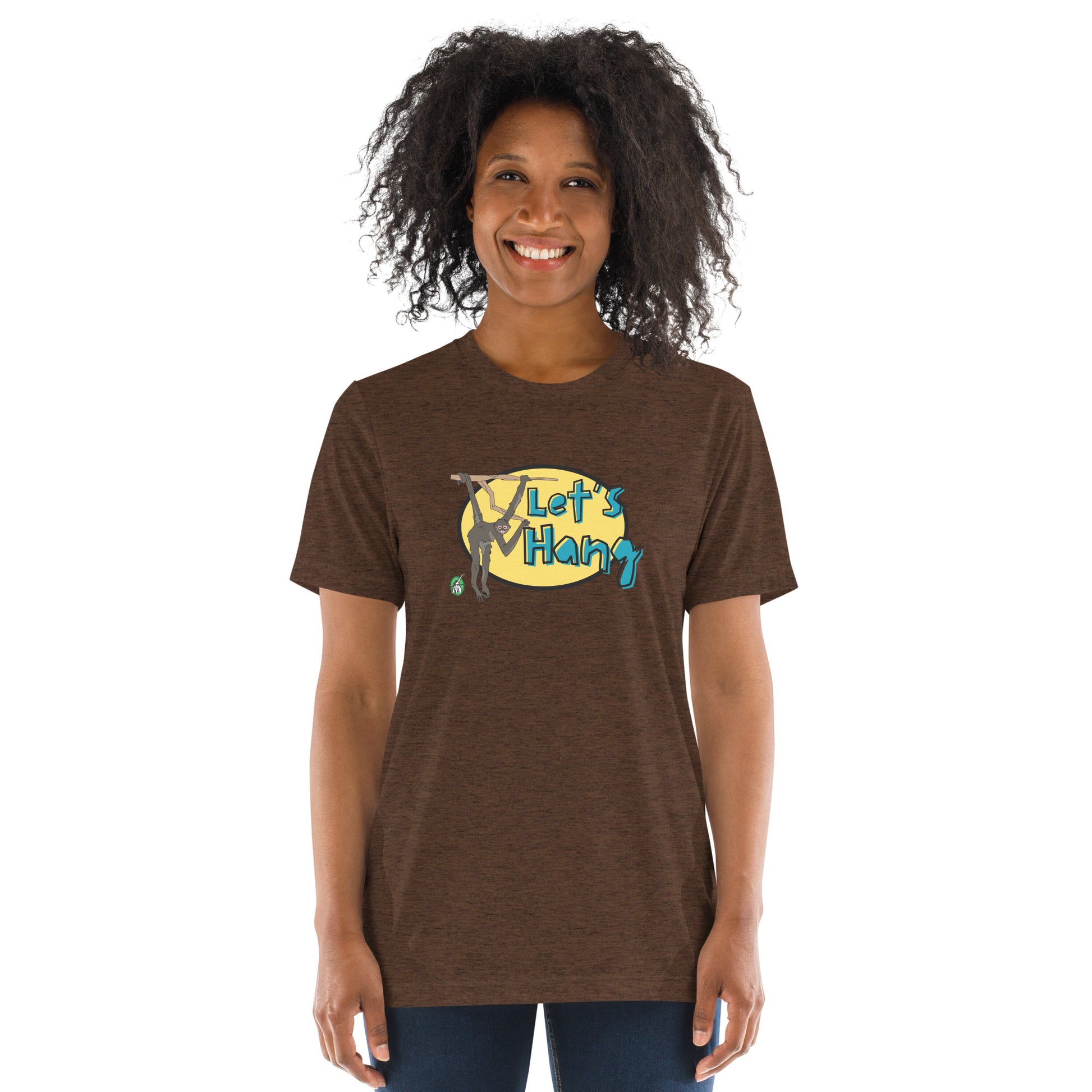 A woman standing wearing a brown tee shirt with a printed graphic of a monkey hanging on a branch with the words, Let's Hang. Designed by Wizard Prints