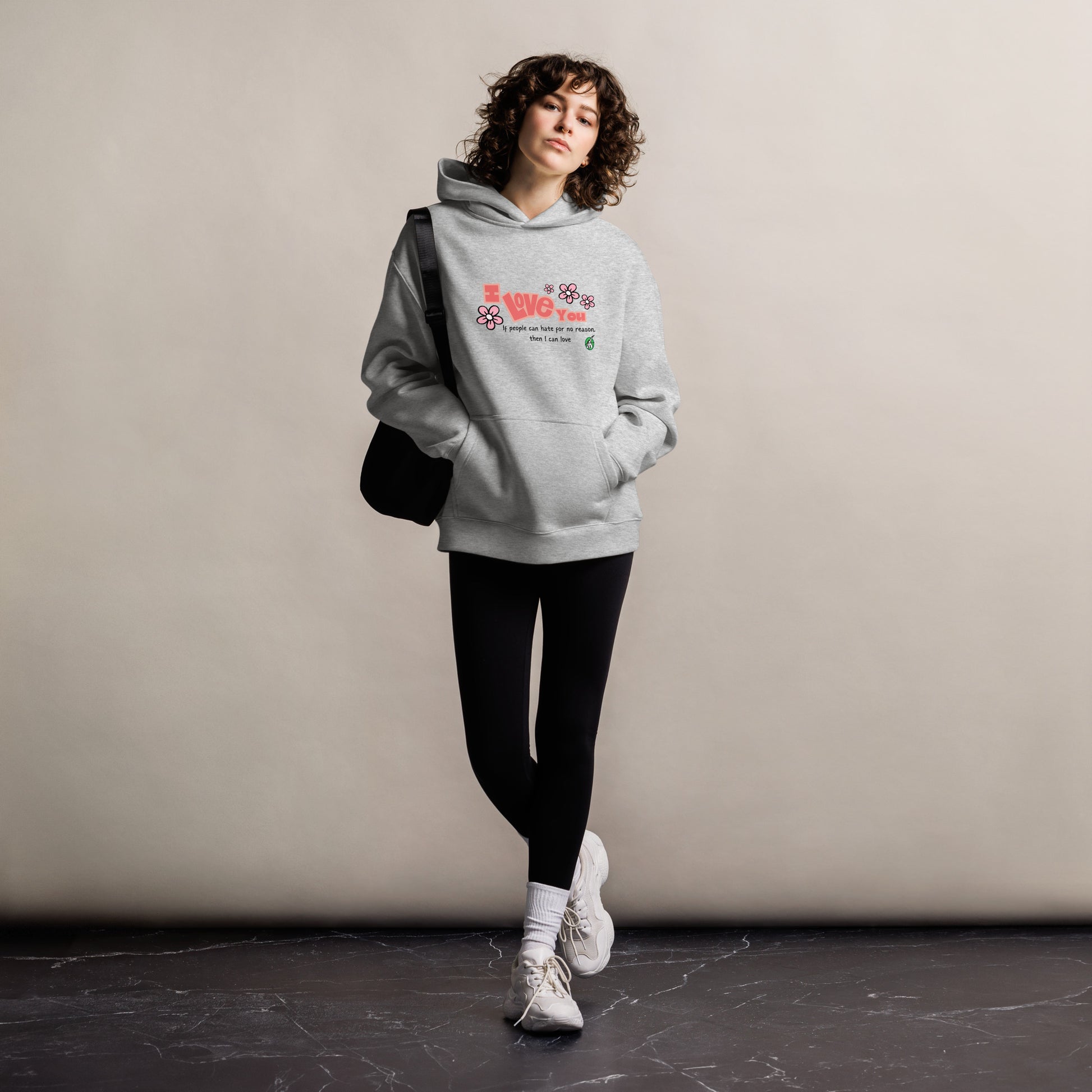 A woman standing wearing a grey hoodie by Wizard Prints featuring the bold text 'I Love You' and the plain text 'If people can hate for no reason, then I can love.'