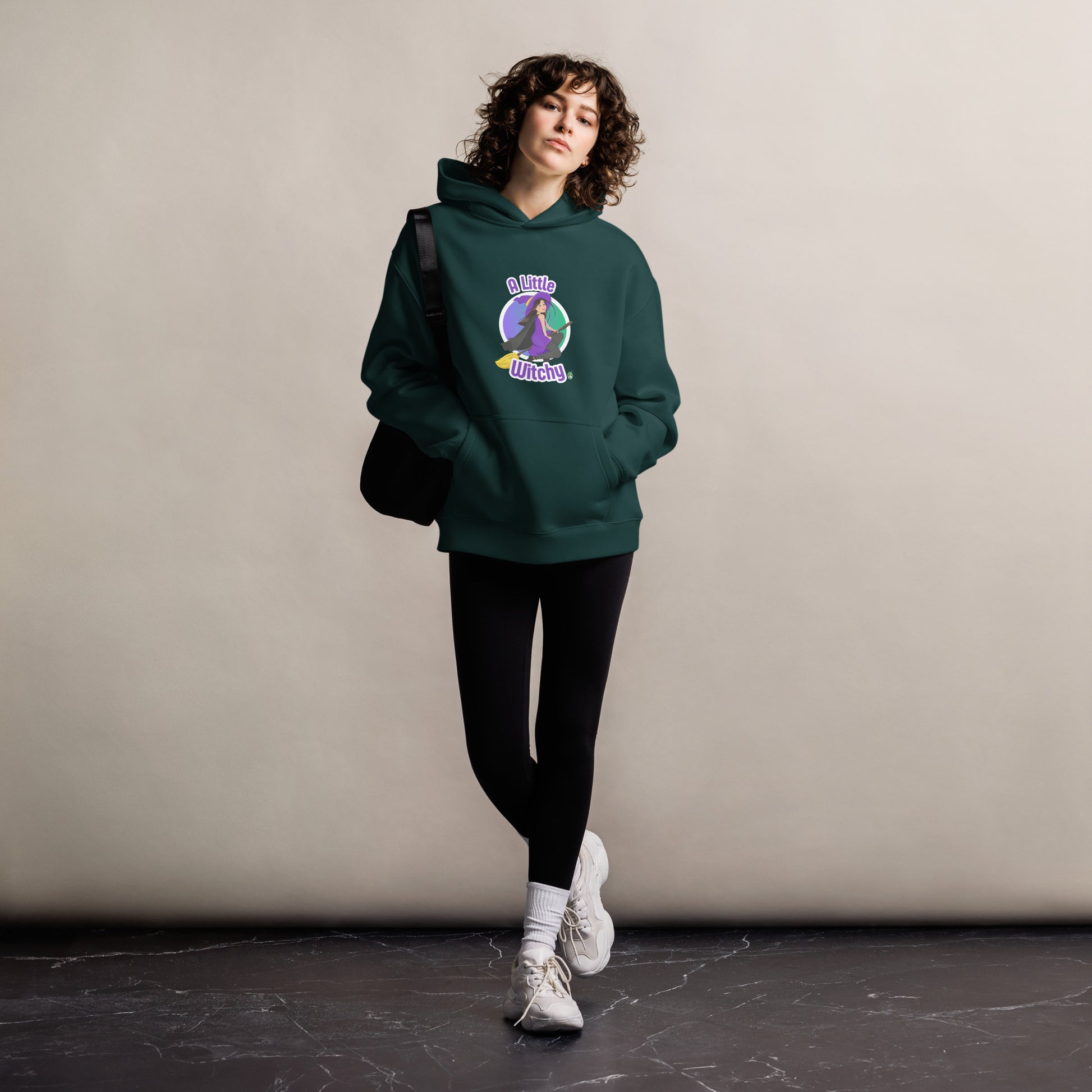 A woman standing wearing a pine green hoodie by Wizard Prints featuring a printed graphic of a witch riding a flying broom, with the text, A Little Witchy.