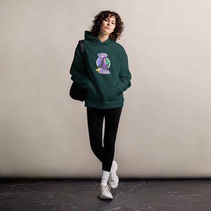 A woman standing wearing a pine green hoodie by Wizard Prints featuring a printed graphic of a witch riding a flying broom, with the text, A Little Witchy.