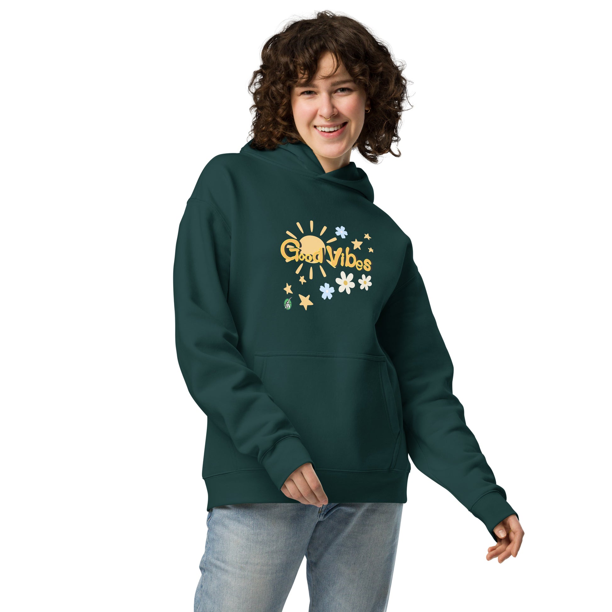 A woman standing wearing a pine green hoodie by Wizard Prints featuring a printed graphic of the sun, flowers, stars and the words, 'Good Vibes'.