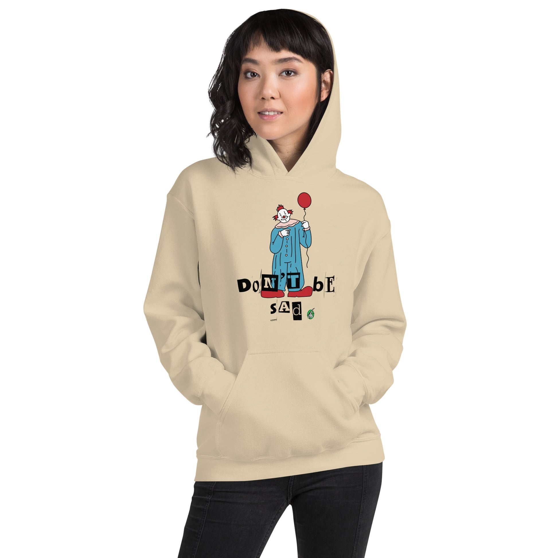 A woman standing and wearing a sand beige hoodie with a printed graphic of a clown holding a balloon with the words, 'Don't Be Sad'. Designed by Wizard Prints.