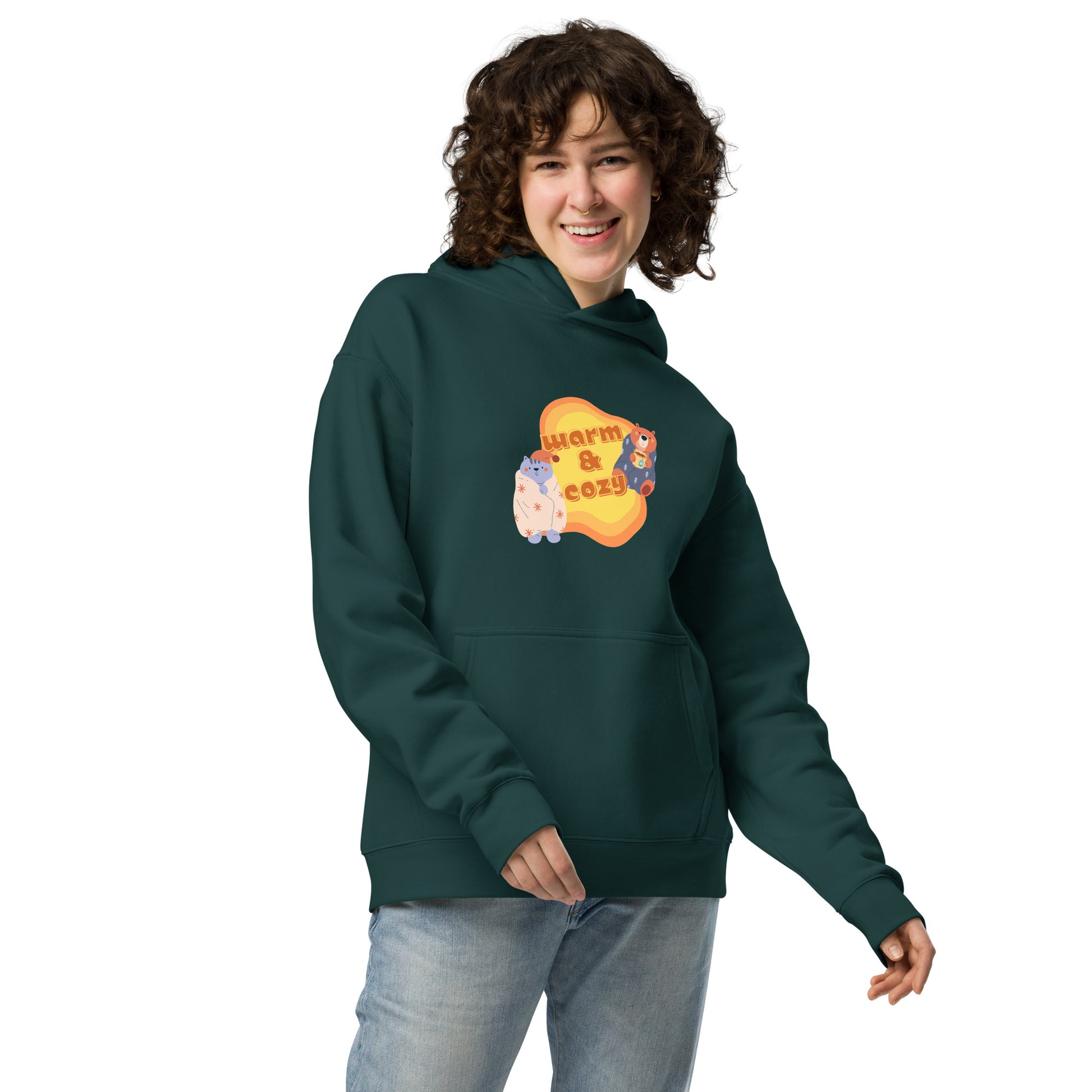 A woman standing wearing a pine green hoodie by Wizard Prints featuring a printed graphic of a cartoon cat and bear wrapped in blankets and the words, Warm & Cozy. 
