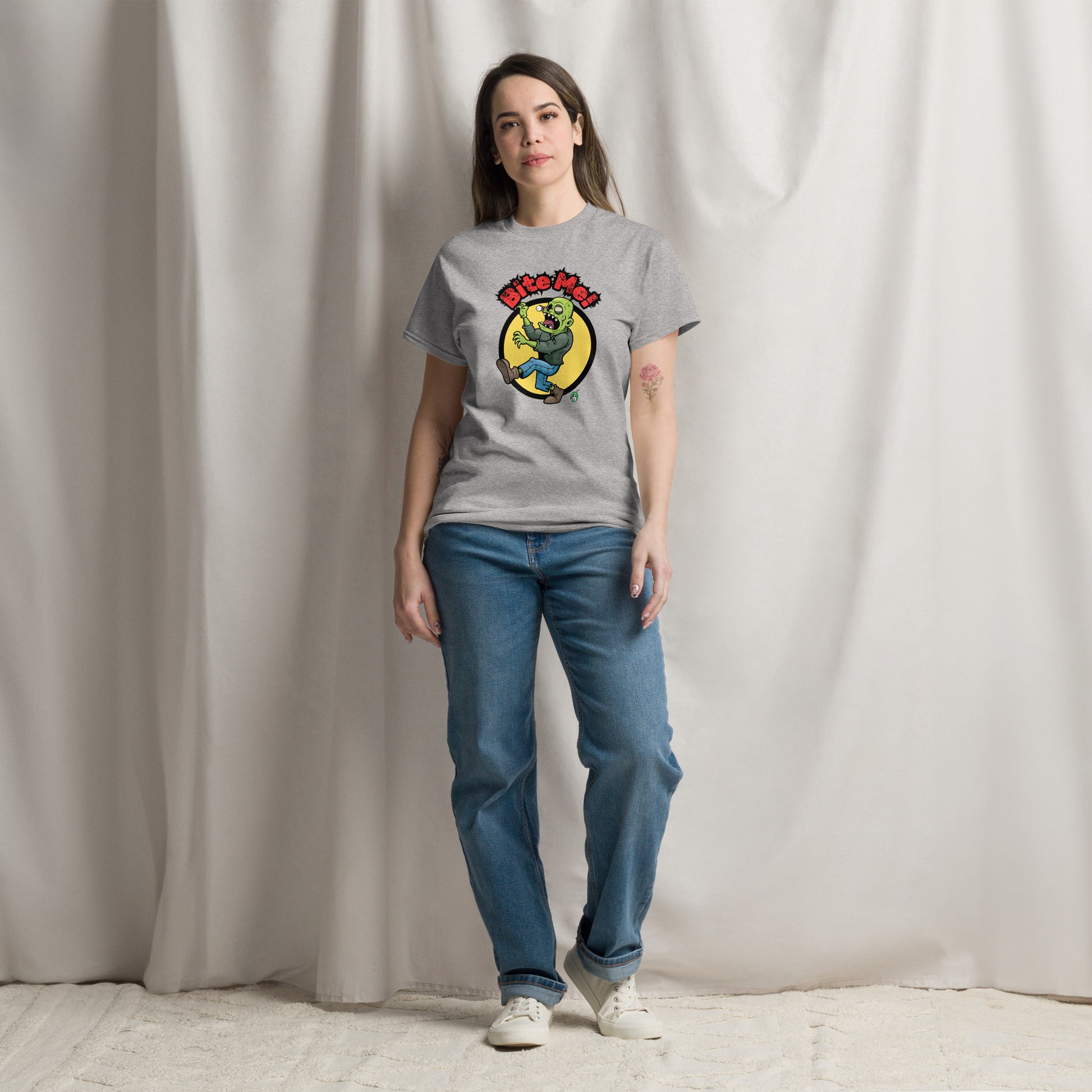 A woman standing and wearing a light grey t-shirt with a printed graphic of a zombie with the words, Bite Me'. Designed by Wizard Prints.