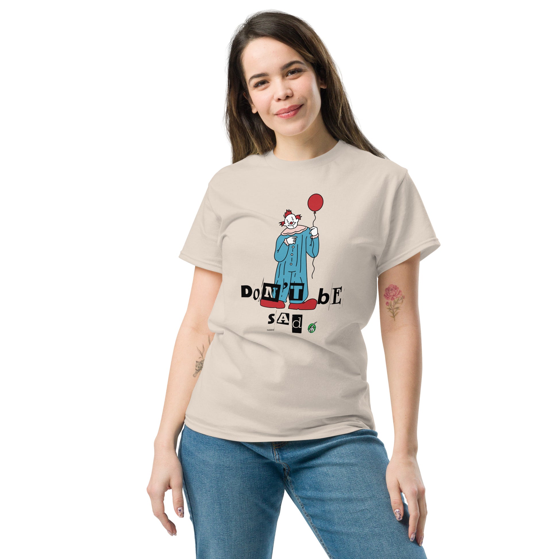 A woman standing and wearing a beige t-shirt with a printed graphic of a clown holding a balloon with the words, "Don't be sad". Designed by Wizard Prints
